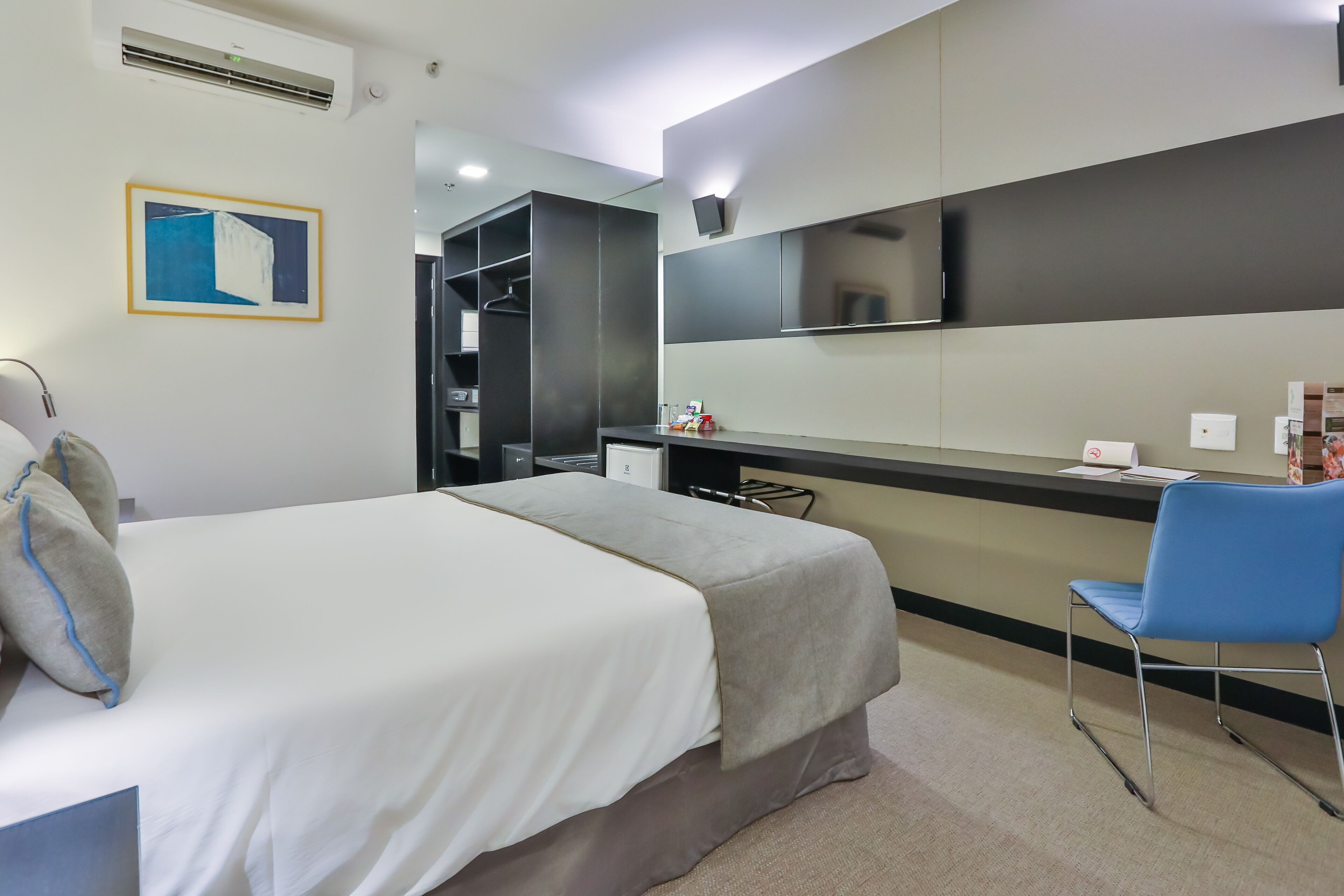 A modern hotel room with a bed, a desk, a chair, a wardrobe, and a flat-screen TV mounted on the wall.