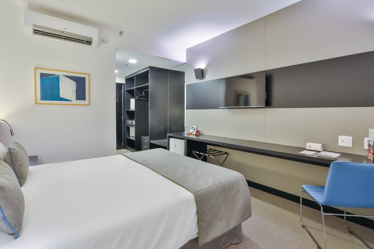 Habitación de hotel con cama, escritorio, silla, armario, espejo y un cuadro en la pared.