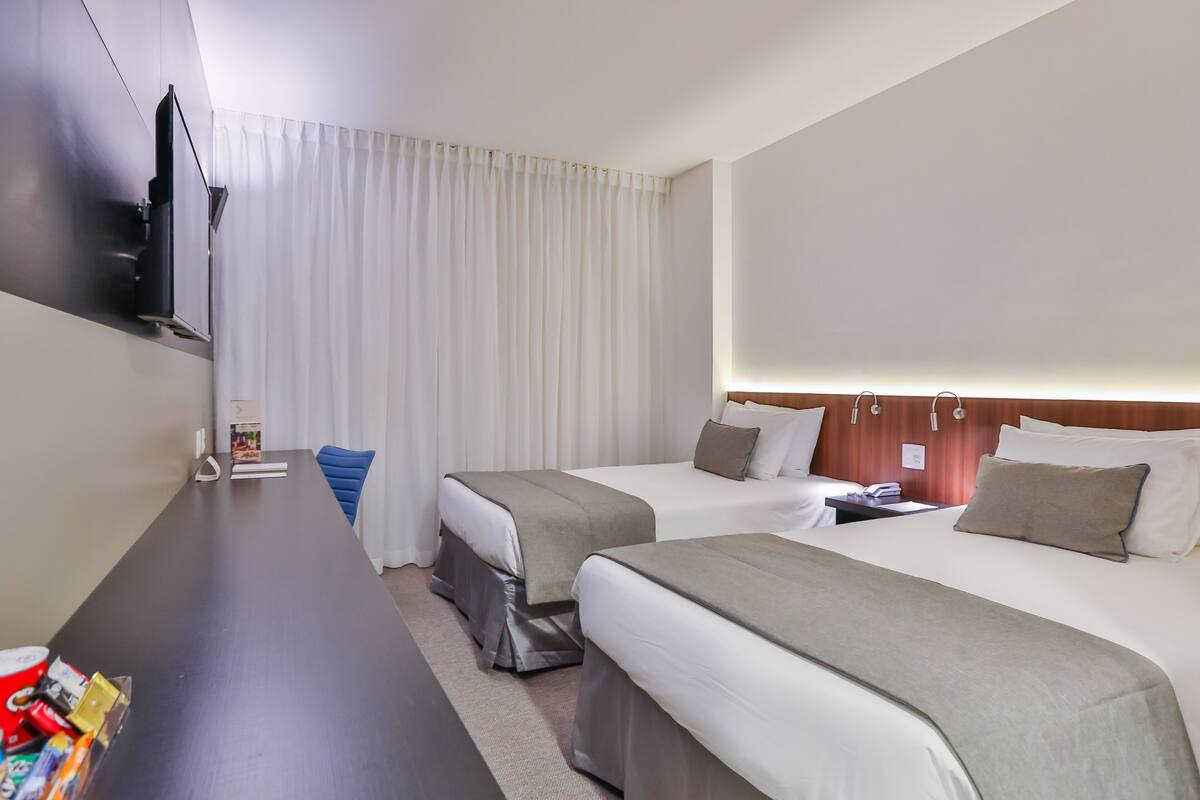 Habitación de hotel con dos camas, un televisor montado en la pared, un escritorio con una silla y un ventanal con cortinas.