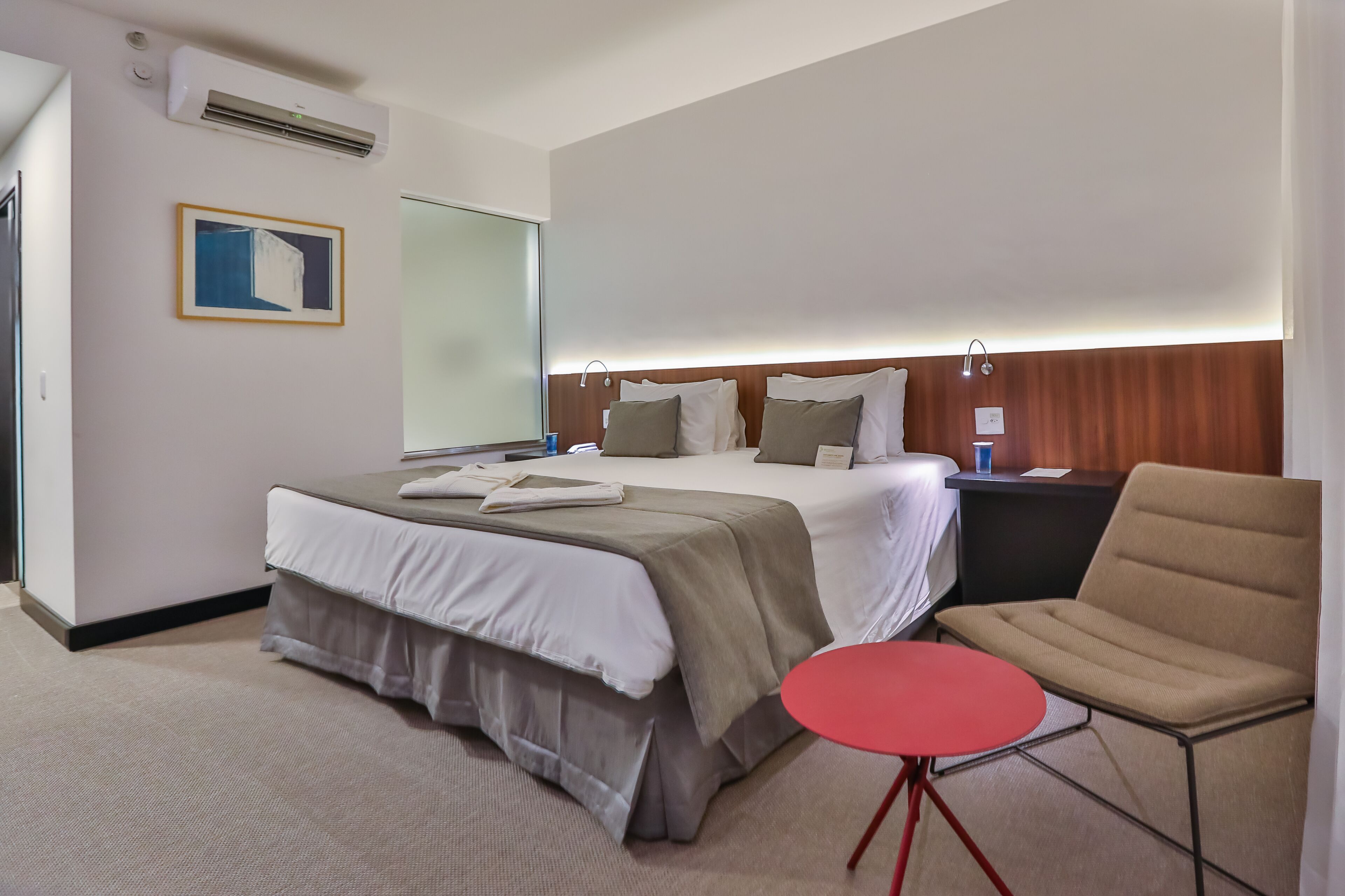 A hotel room with a large bed, a bedside table, a red chair, and a small round table.