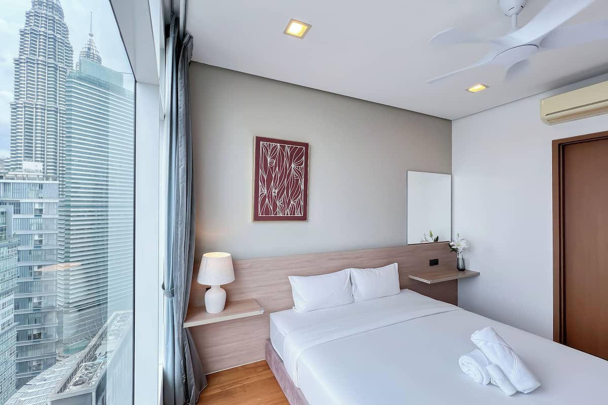A modern hotel room with a large bed, a bedside table with a lamp, a small shelf, a mirror, and a view of a cityscape.