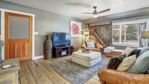 Una sala de estar con piso de madera, televisor, chimenea, ventilador de techo y un amplio ventanal con cortinas.
