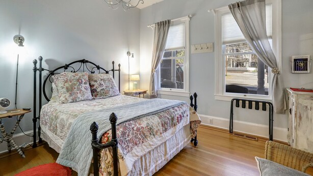 A bedroom with a bed, a nightstand, a lamp, a radiator, and a window with curtains. - Broad Street Inn (Nevada City)
