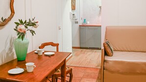 Un comedor con una mesa de madera, sillas y un florero. Se ve un sofá y una cocina al fondo.
