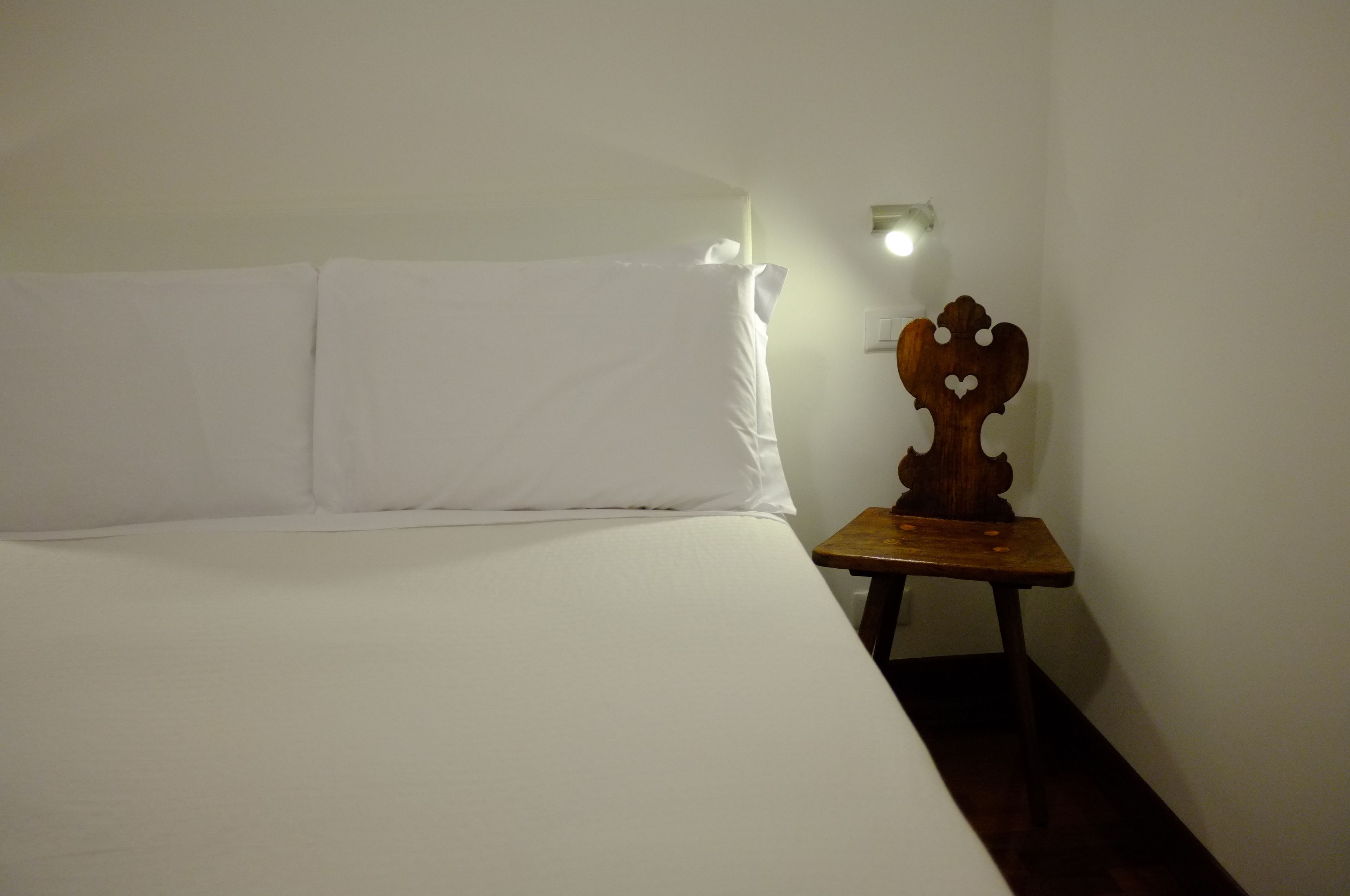 Ein Einzelbett mit weißer Bettwäsche in einem Zimmer mit einem kleinen Nachttisch aus Holz und einer wandmontierten Lampe.
