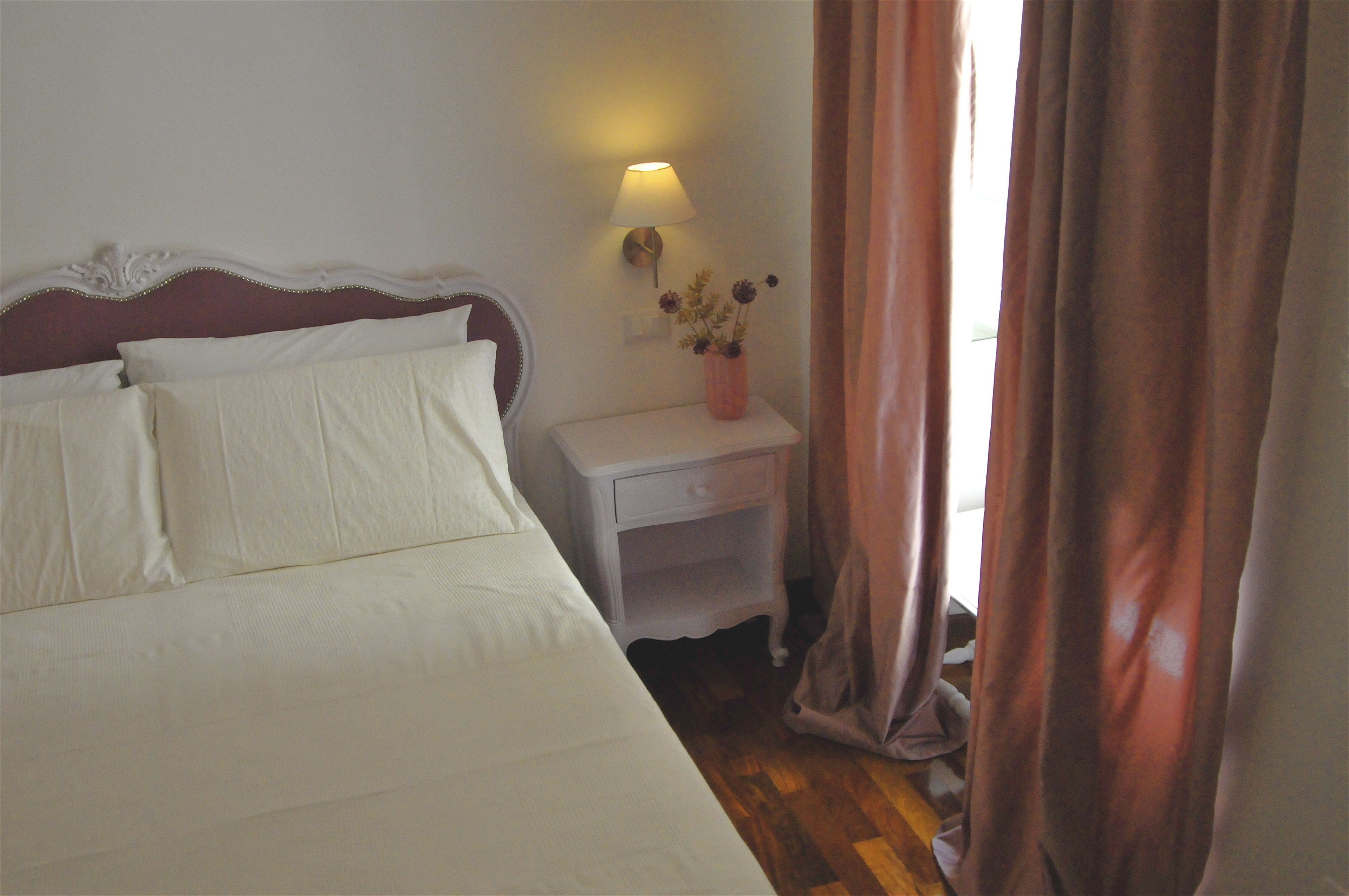A neatly made bed with white linens, a white nightstand with a lamp and a vase of flowers, and a wooden floor.