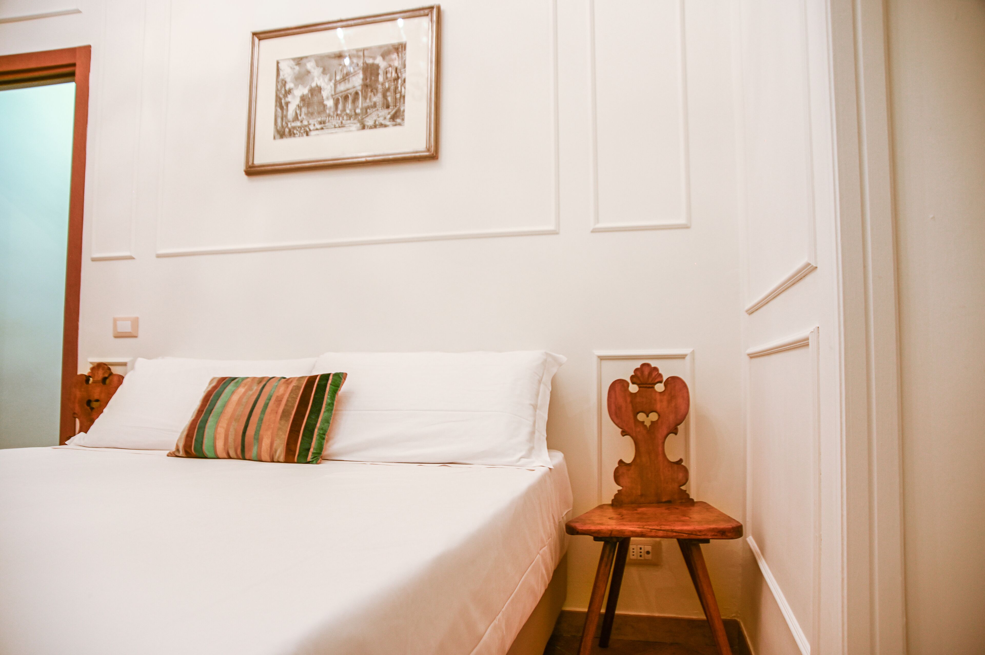 Una cama bien hecha con sábanas blancas y una almohada a rayas, una silla de madera con respaldo en forma de corazón y un cuadro en la pared.