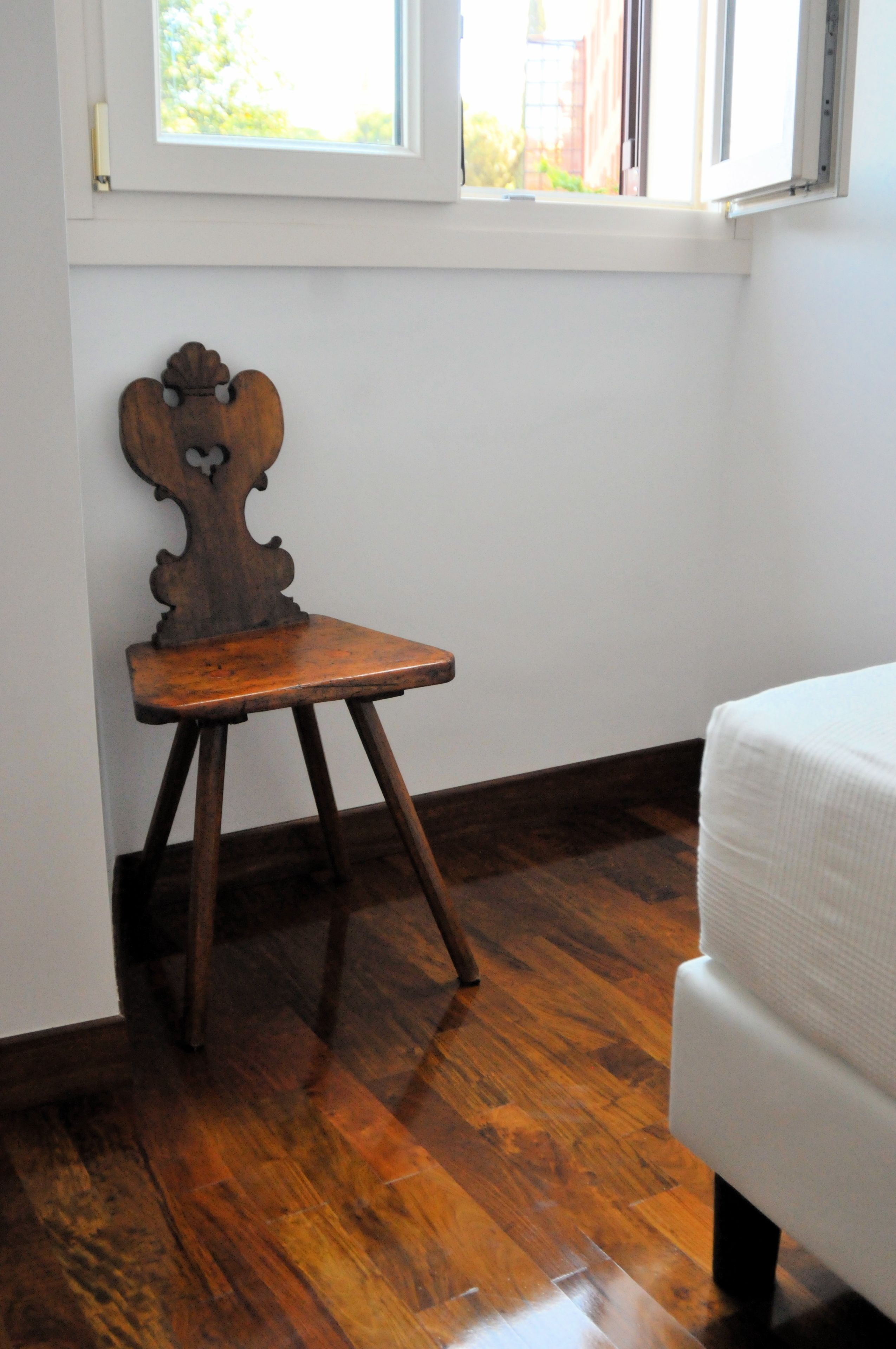 A room with a wooden chair against a wall, a white sofa, and a window with a view of greenery outside.