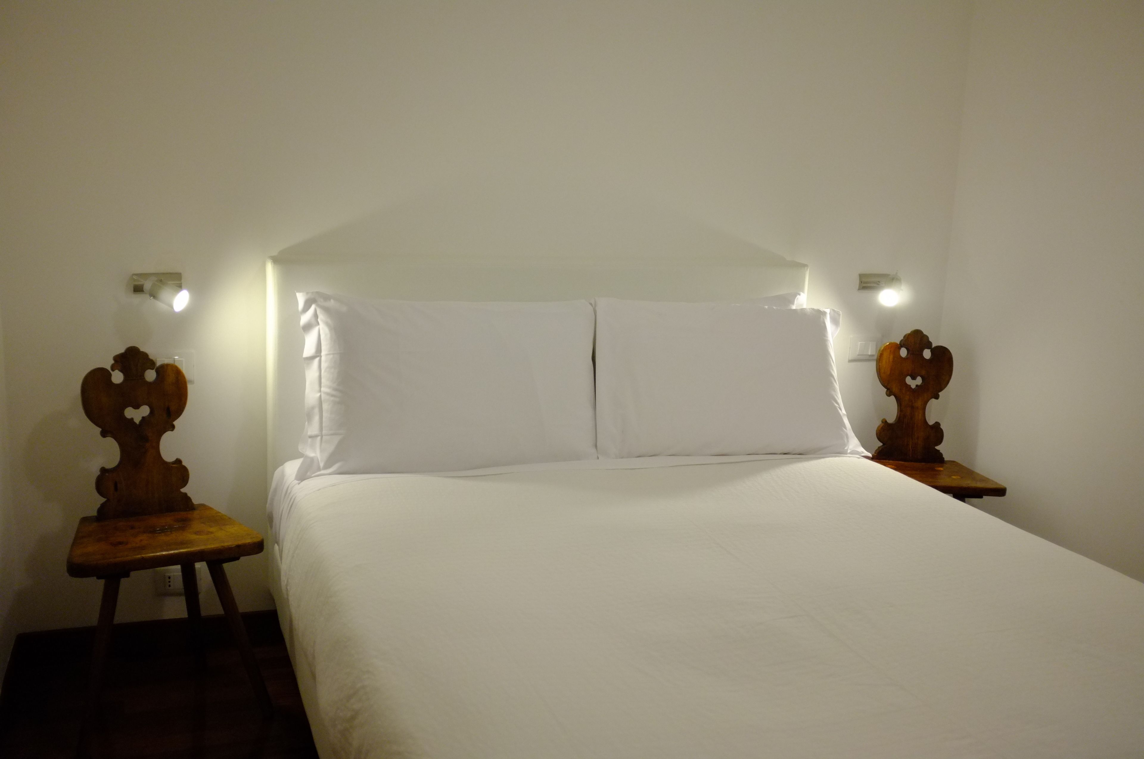 Un lit simple avec du linge de lit blanc, dans une pièce aux murs blancs et au plafond en pente. On y trouve deux tables de chevet en bois, ornées de plaques décoratives, et une petite table placée entre les lits.