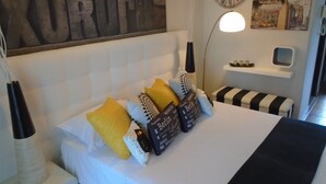 Una cama bien hecha con cabecera blanca, varios almohadones, una mesita de noche con lámpara, un reloj y un cuadro en la pared.