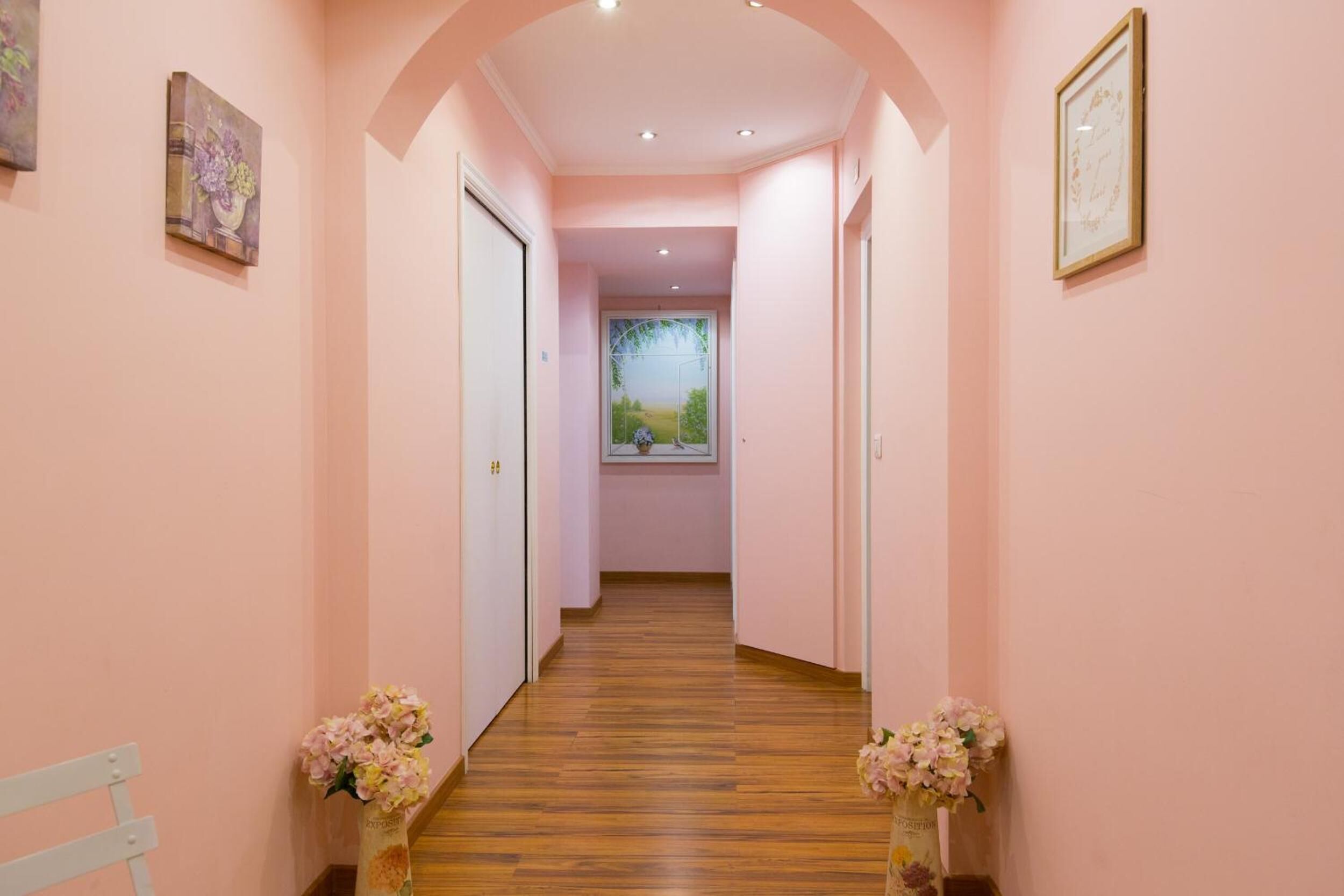 A hallway with pink walls, wooden flooring, and two vases with flowers at the ends.
