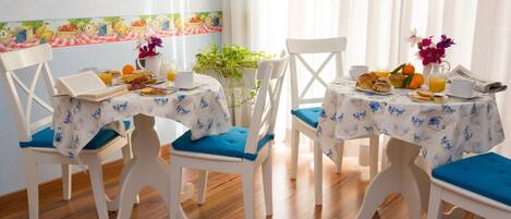 A breakfast setting with two white chairs, a table with a floral tablecloth, and a variety of breakfast items including croissants, oranges, and a pitcher of orange juice.