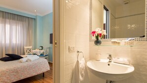 Una habitación de hotel con cama, baño con lavamanos y espejo, y una mesita pequeña con silla.
