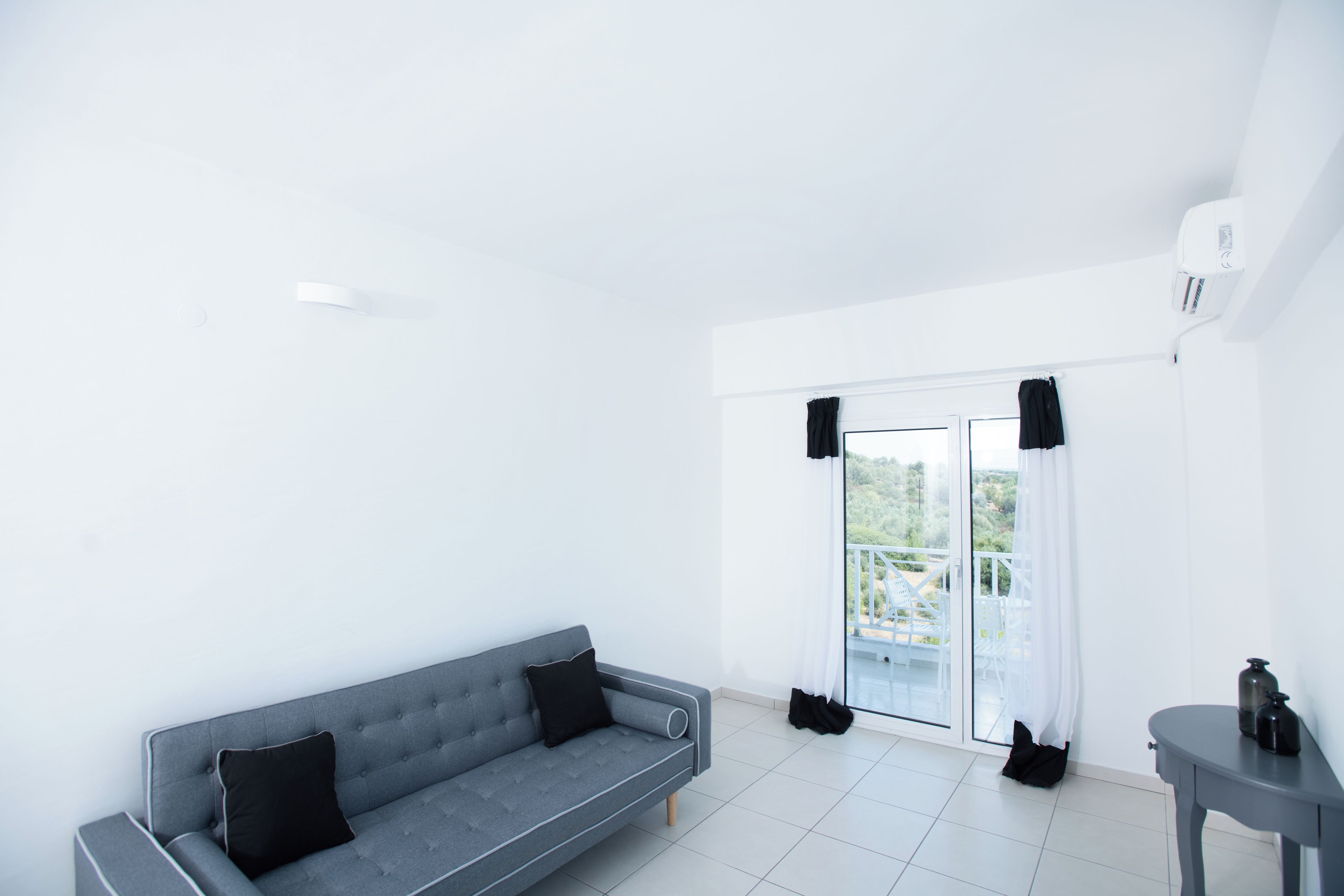 A modern, minimalist room with a grey sofa, a small table, and a sliding glass door leading to an outdoor balcony.