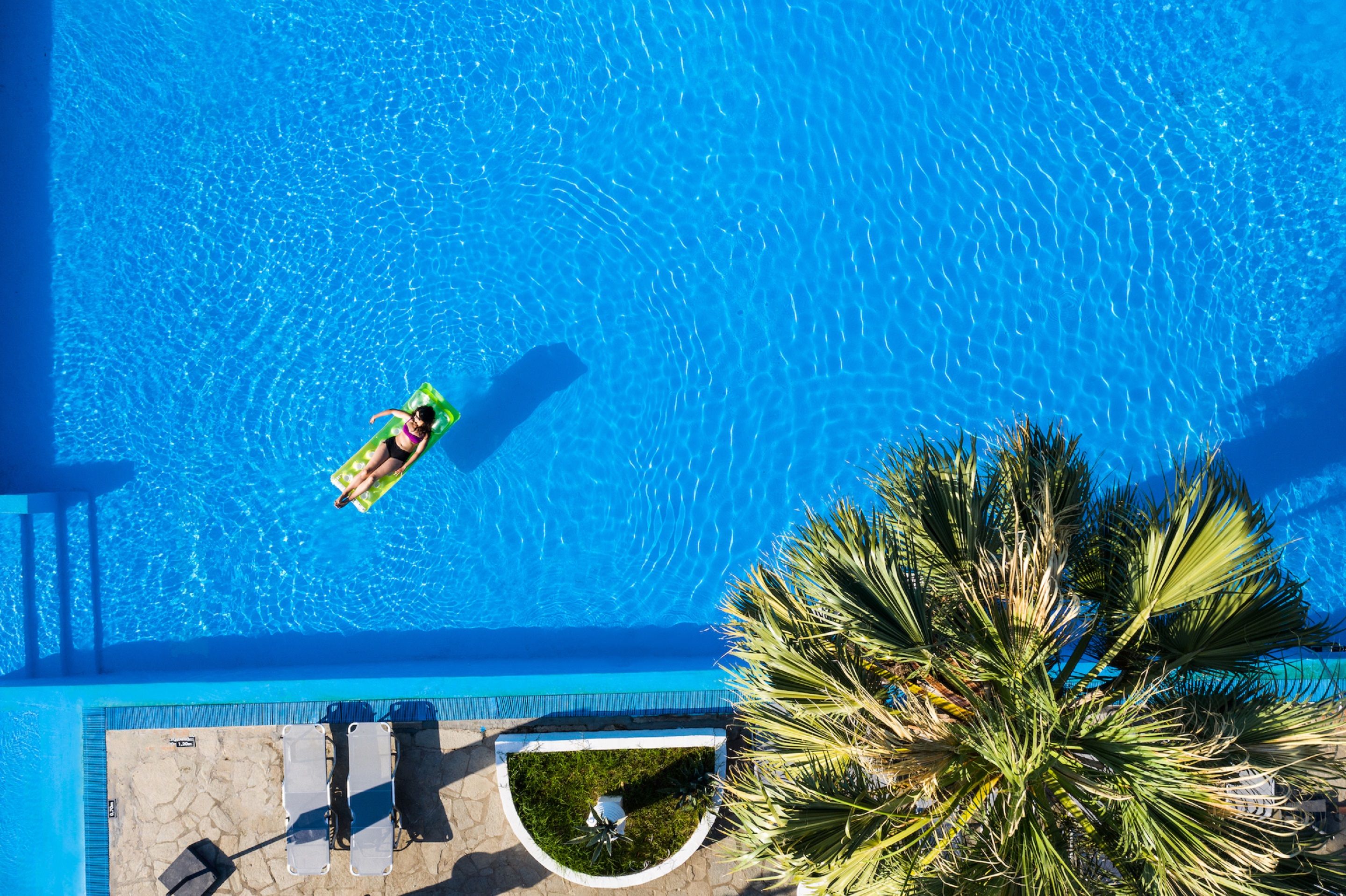 Eine Vogelperspektive zeigt einen Swimmingpool mit einer Person auf einem Schwimmschlauch, Palmen und Liegestühlen.