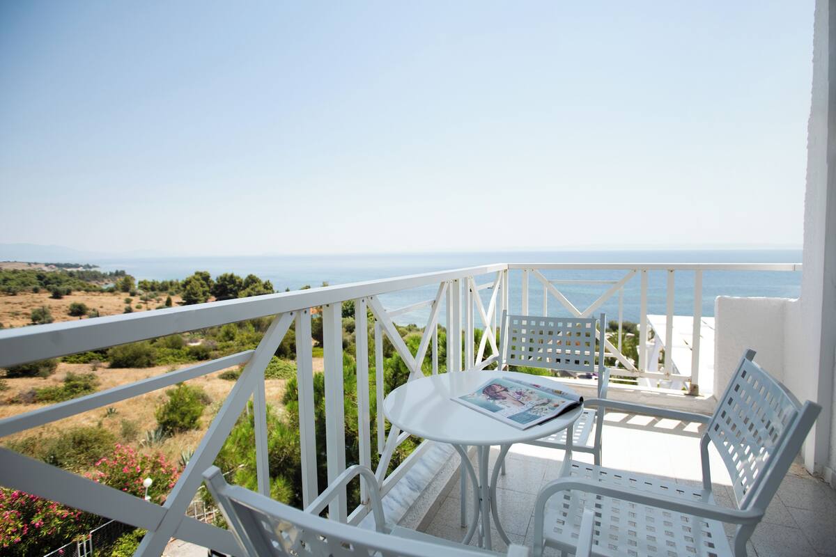 A balcony with white metal railings, a small round table with a magazine, and two white metal chairs. The balcony overlooks a coastal landscape with greenery and a clear sky.