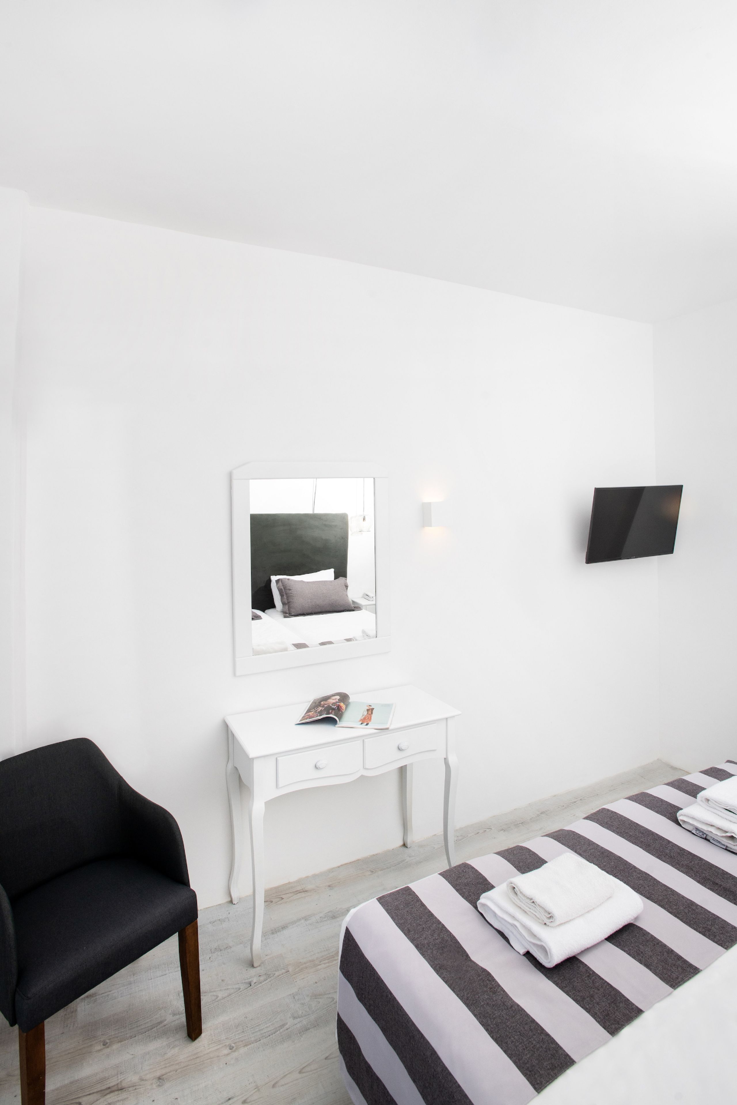 A minimalist hotel room with a bed, a white desk, a black chair, a flat-screen TV, and a mirror.