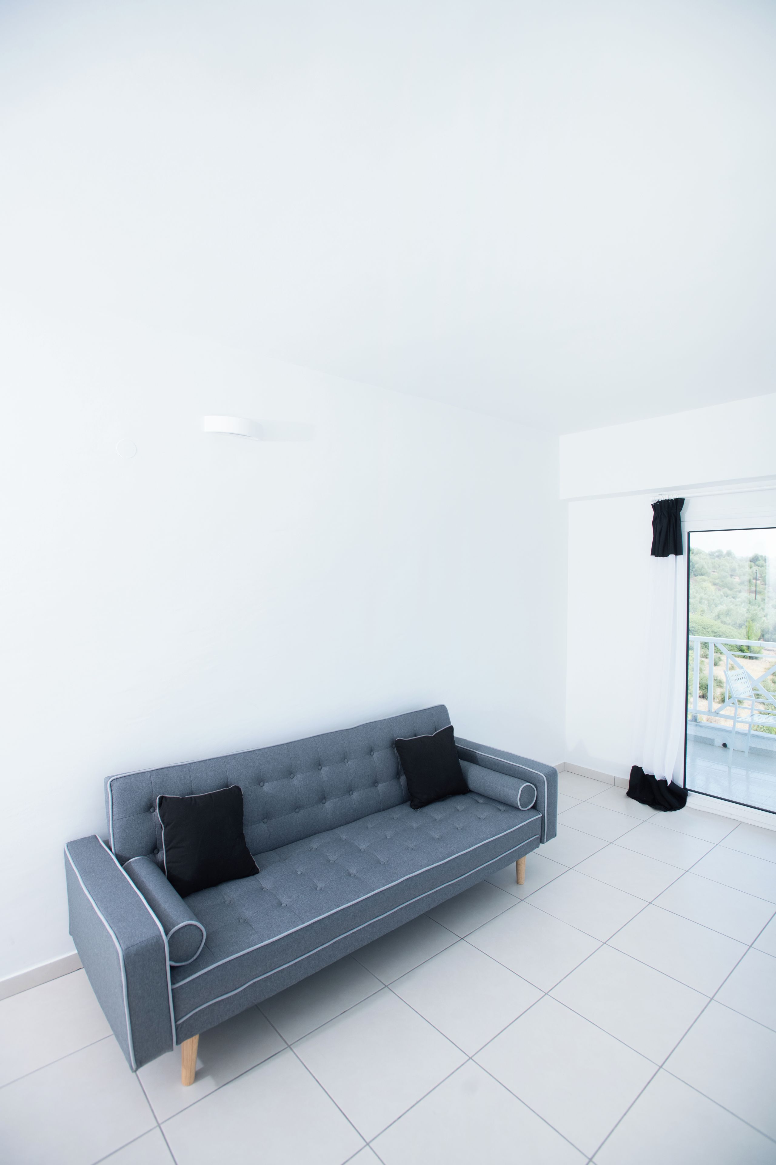 A modern, grey sofa with two black cushions in a bright, minimally furnished room with white walls and a large window.