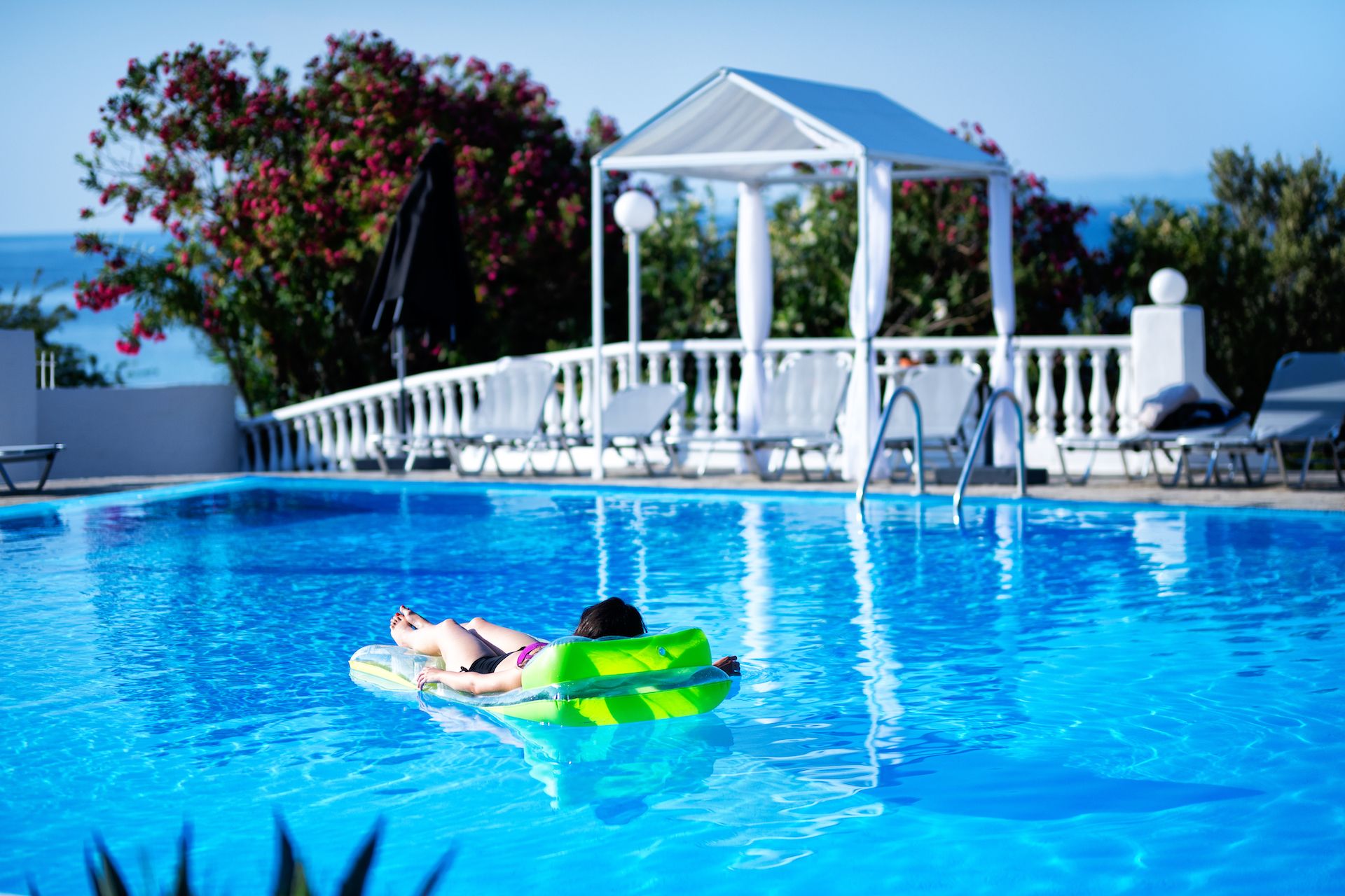 A swimming pool with a person on a green float, lounge chairs, and a gazebo in the background.