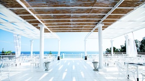 An outdoor terrace with white chairs and tables, a wooden ceiling, and a view of the sea. - Bianco Olympico Beach Resort By Anayia AI Resorts (Polygyros)