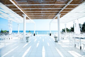 An outdoor terrace with white chairs and tables, a wooden ceiling, and a view of the sea.