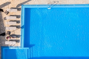 A rectangular swimming pool with a clear blue water surface, surrounded by a stone-paved deck with several lounge chairs and umbrellas.