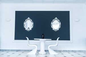 Una habitación con una pared azul oscuro de acento, dos espejos blancos ornamentados, una mesa blanca con una botella negra y sillas blancas.