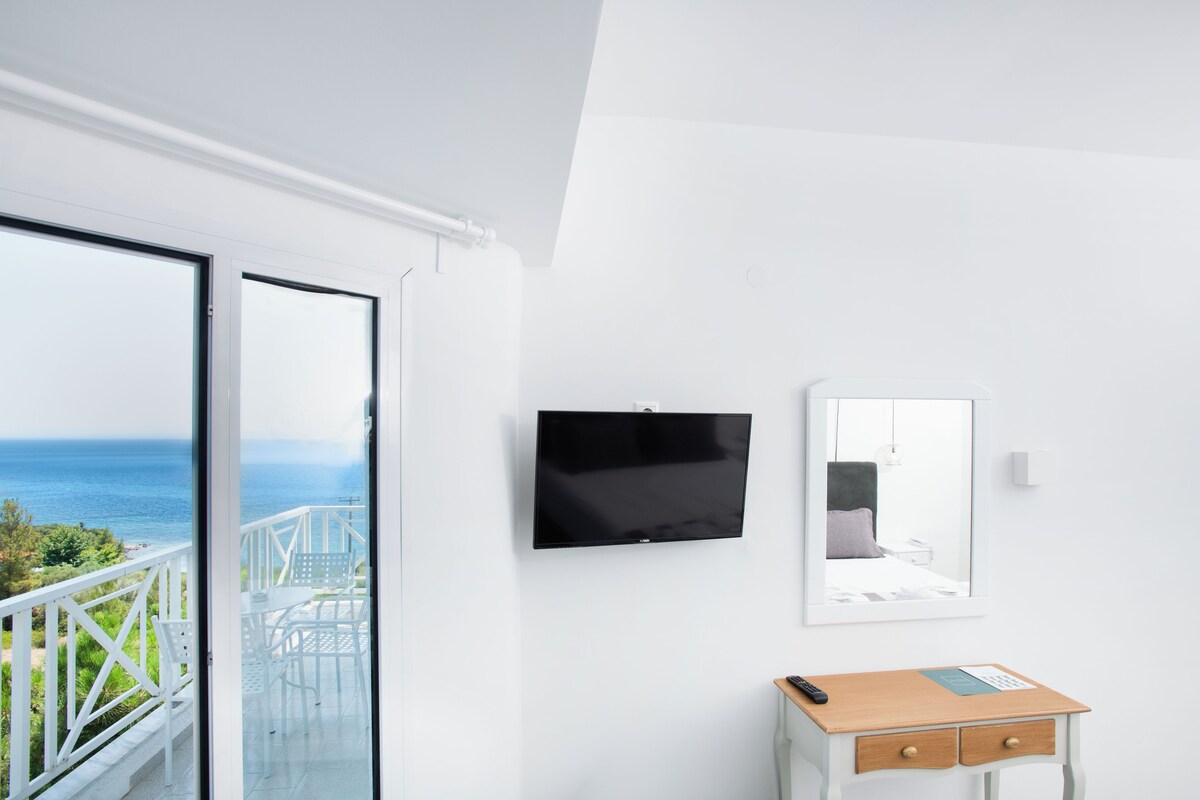 Una habitación con televisor montado en la pared, un escritorio pequeño con cajones y un balcón con vistas al mar.
