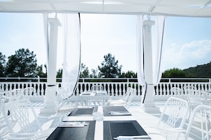 Un área de comedor al aire libre con sillas y mesas blancas, barandal blanco y vista a árboles y colinas.