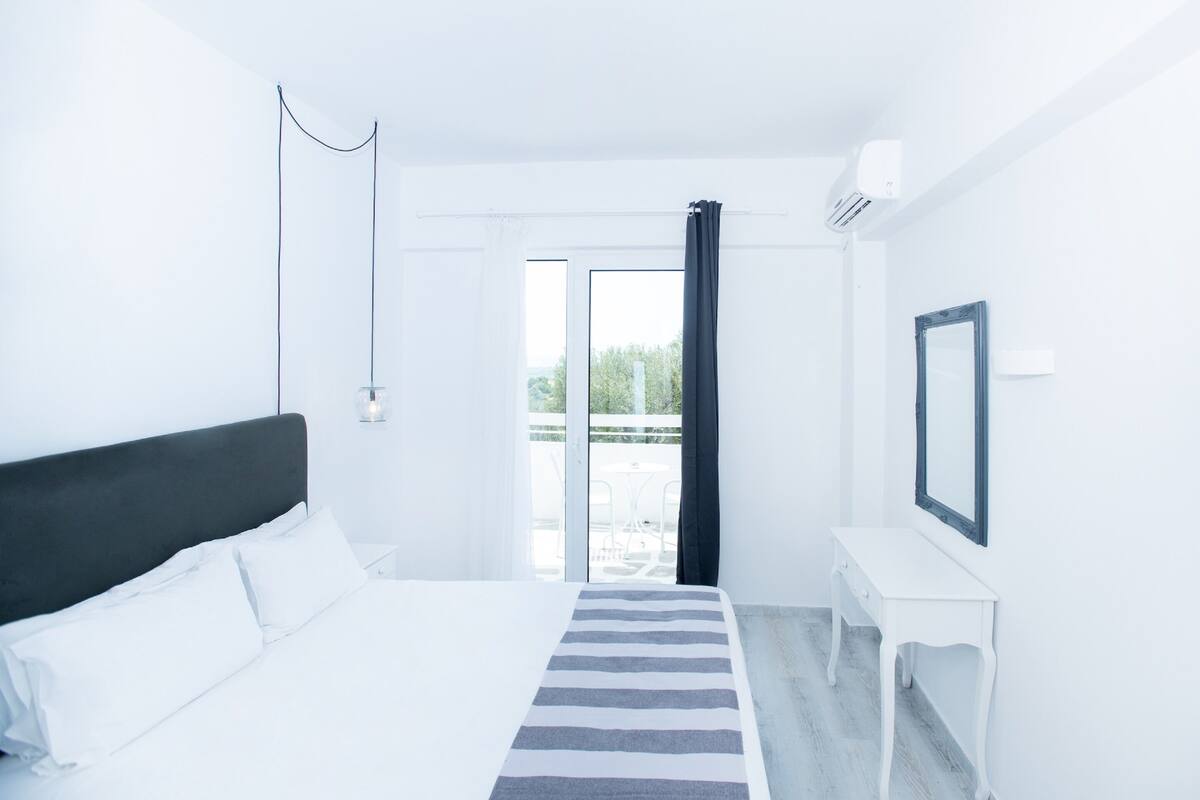 A hotel room with a bed, a bedside table, a mirror, and a view of the outdoors.