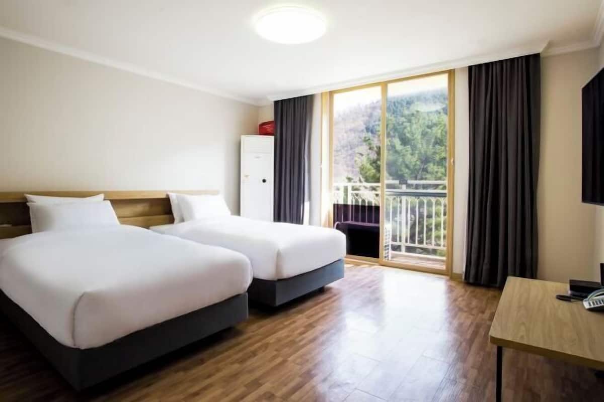 A hotel room with two beds, a wooden floor, a desk with a chair, a wardrobe, and a balcony with a view of greenery.