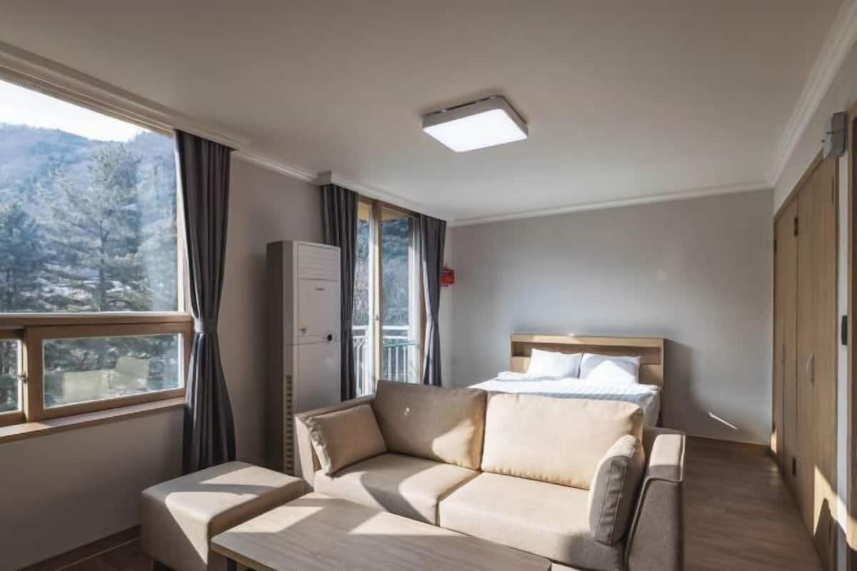 A bedroom with a bed, a sofa, a desk, and a wardrobe. There is a window with curtains and a view of trees.