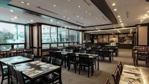 Un restaurante amplio con varias mesas y sillas, ventanas grandes y una zona de cocina al fondo.