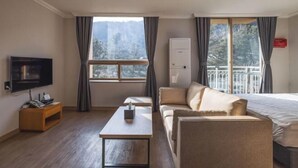 Habitación de hotel con televisor, sofá, mesita, cama y ventana con vista a árboles.