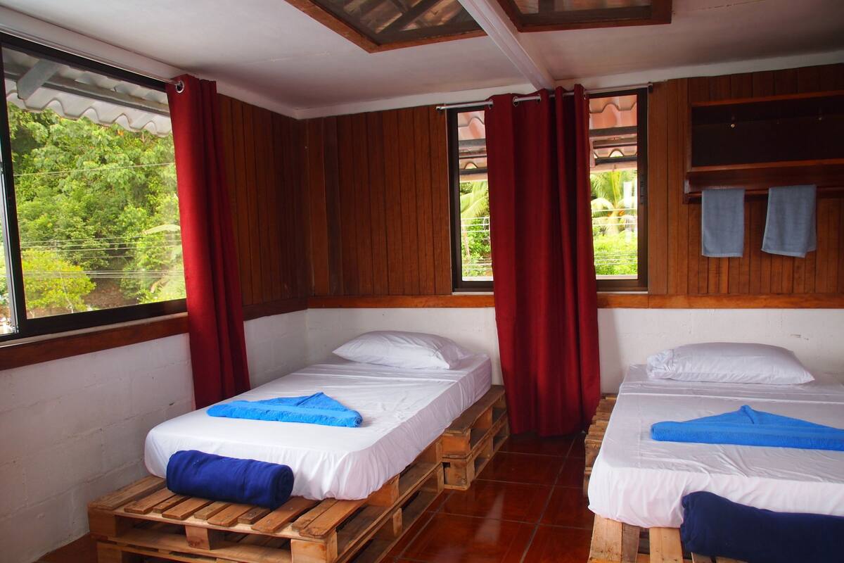 A room with two beds on wooden pallets, red curtains, and a view of greenery outside.