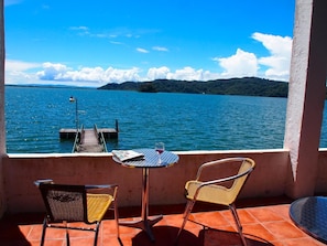 A terrace with two wicker chairs, a small table, and a glass of red wine. A wooden dock extends into a large body of water, with hills in the background. - Hostal del Mar (Golfito)