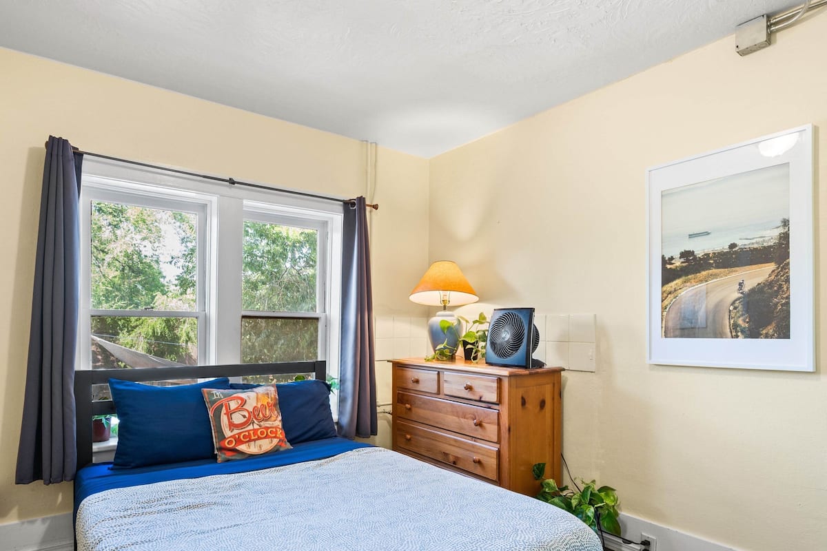 A bedroom with a bed, a dresser, a lamp, a fan, a window with curtains, and a framed picture on the wall.