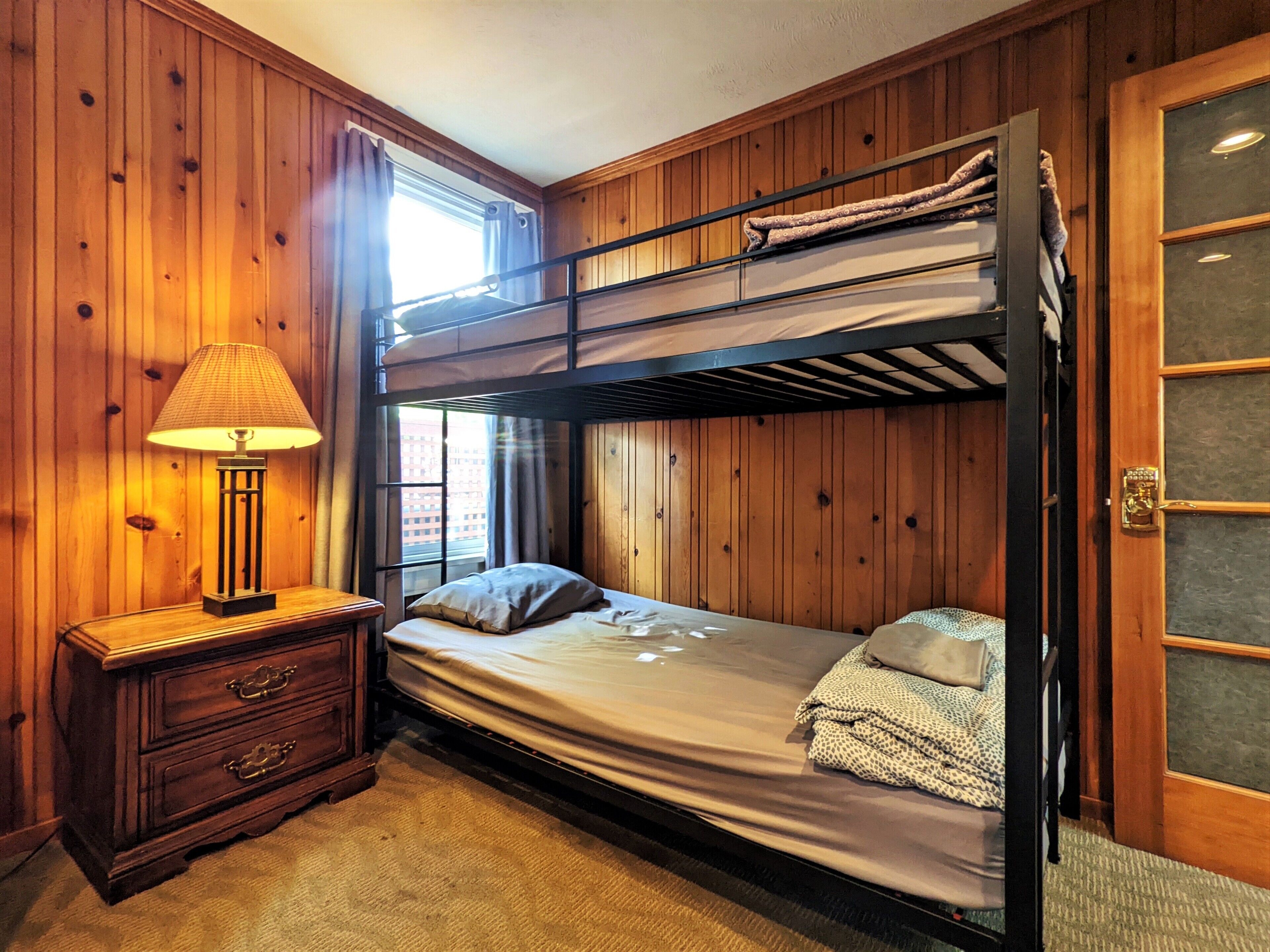 A bunk bed room with a wooden wall, a nightstand with a lamp, and a window with curtains.