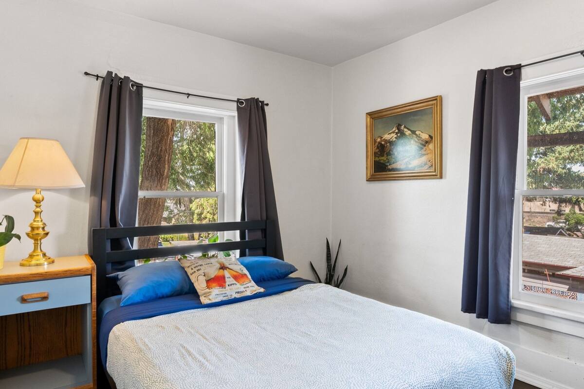A bedroom with a bed, a nightstand, a lamp, a window with curtains, and a painting on the wall.