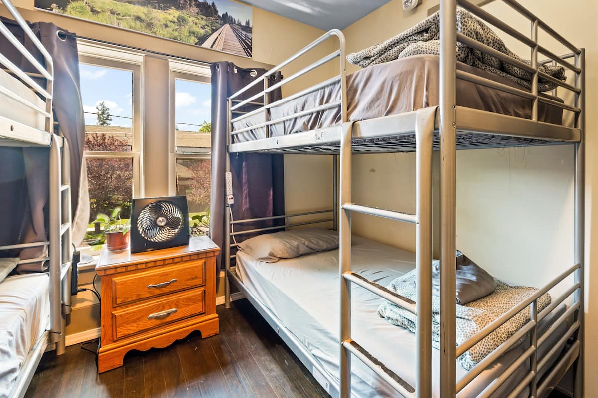 A dormitory room with bunk beds, a wooden dresser, a fan, and a window with a view of greenery.