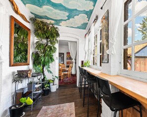 Una habitación acogedora con una mesa de madera, sillas y un mural en el techo. Hay plantas en macetas y una estantería.