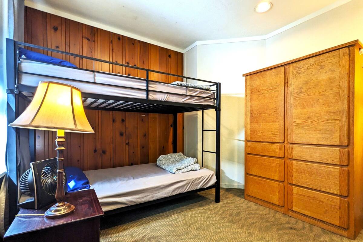 A bunk bed room with a wooden wall, a desk with a lamp, and a wardrobe.
