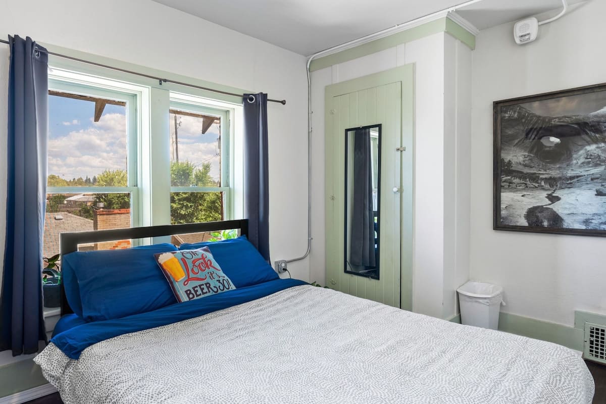 A bedroom with a bed, a window with curtains, a door, a painting, and a trash bin.