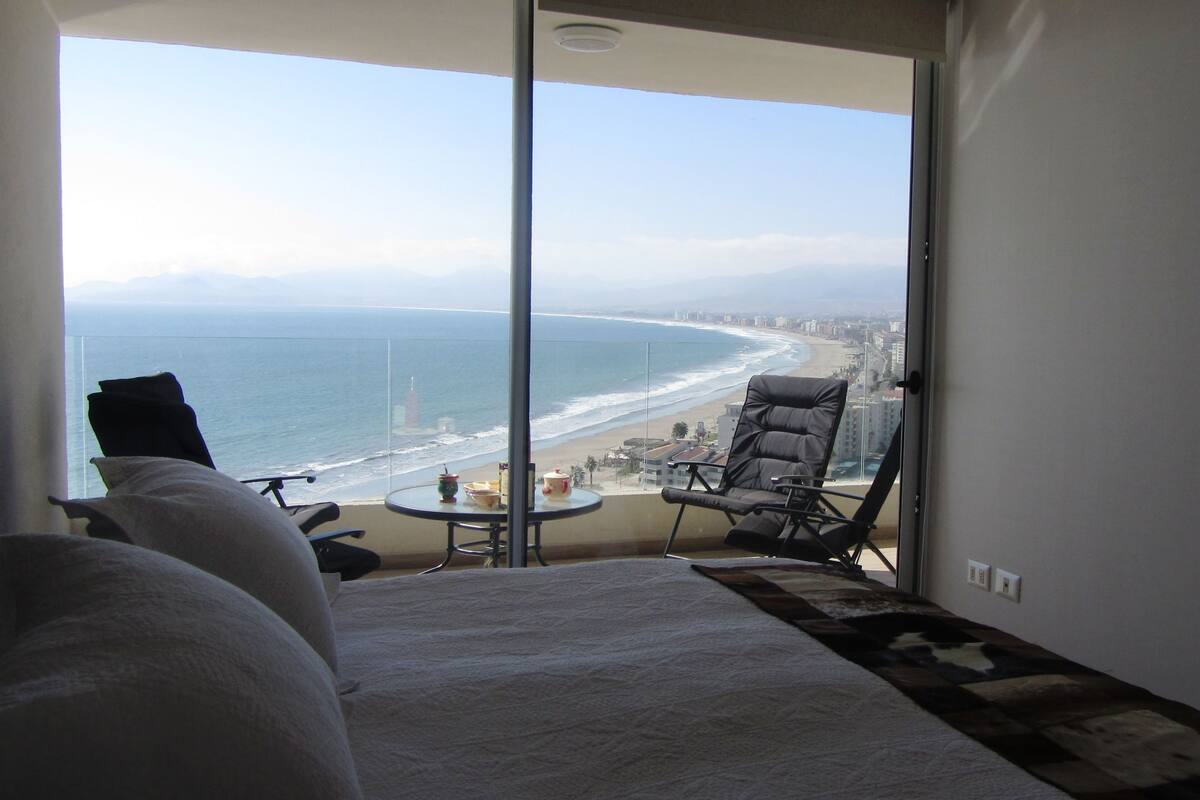 A balcony with a view of the beach and ocean, a chair, a small table with a drink, and a sofa.