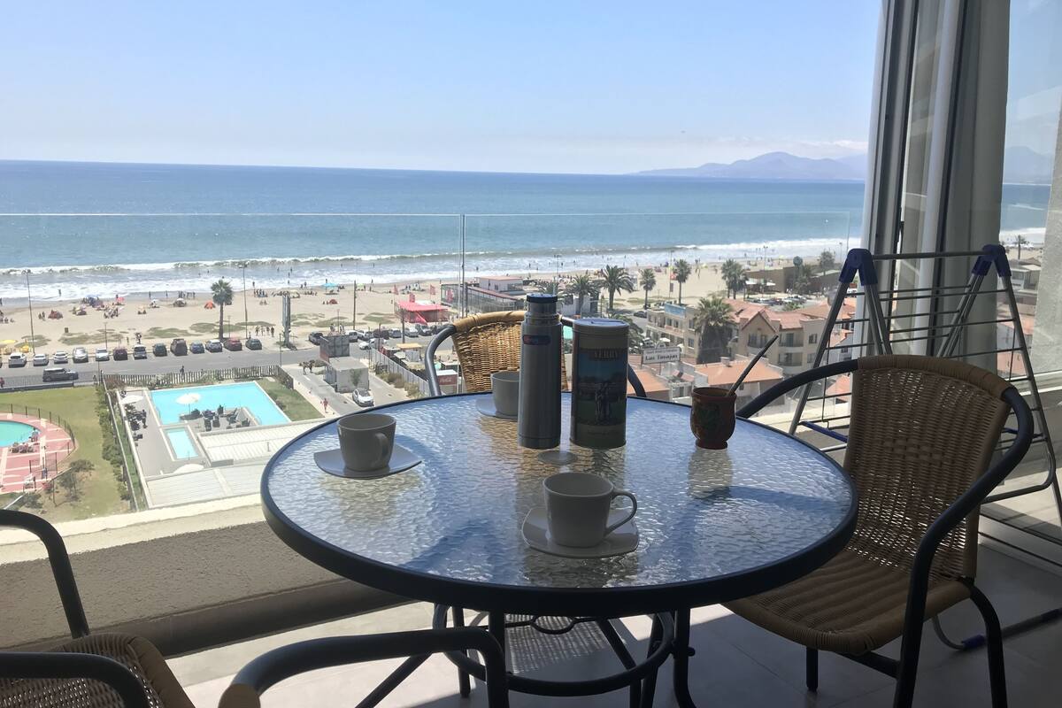 A balcony with a glass table and wicker chairs overlooking a beach and ocean.