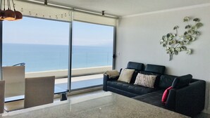 Un salon moderne doté d’une grande fenêtre donnant sur la mer, d’un canapé noir agrémenté de coussins à motifs et d’une plante décorative accrochée au mur.