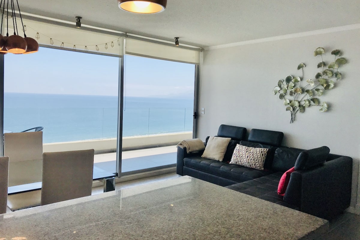 A modern living room with a large window overlooking the sea, a black sofa with patterned cushions, and a decorative plant on the wall.