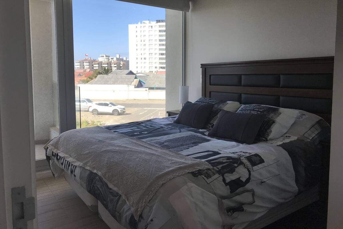 A bedroom with a large bed, a window with a view of buildings and a parking lot.