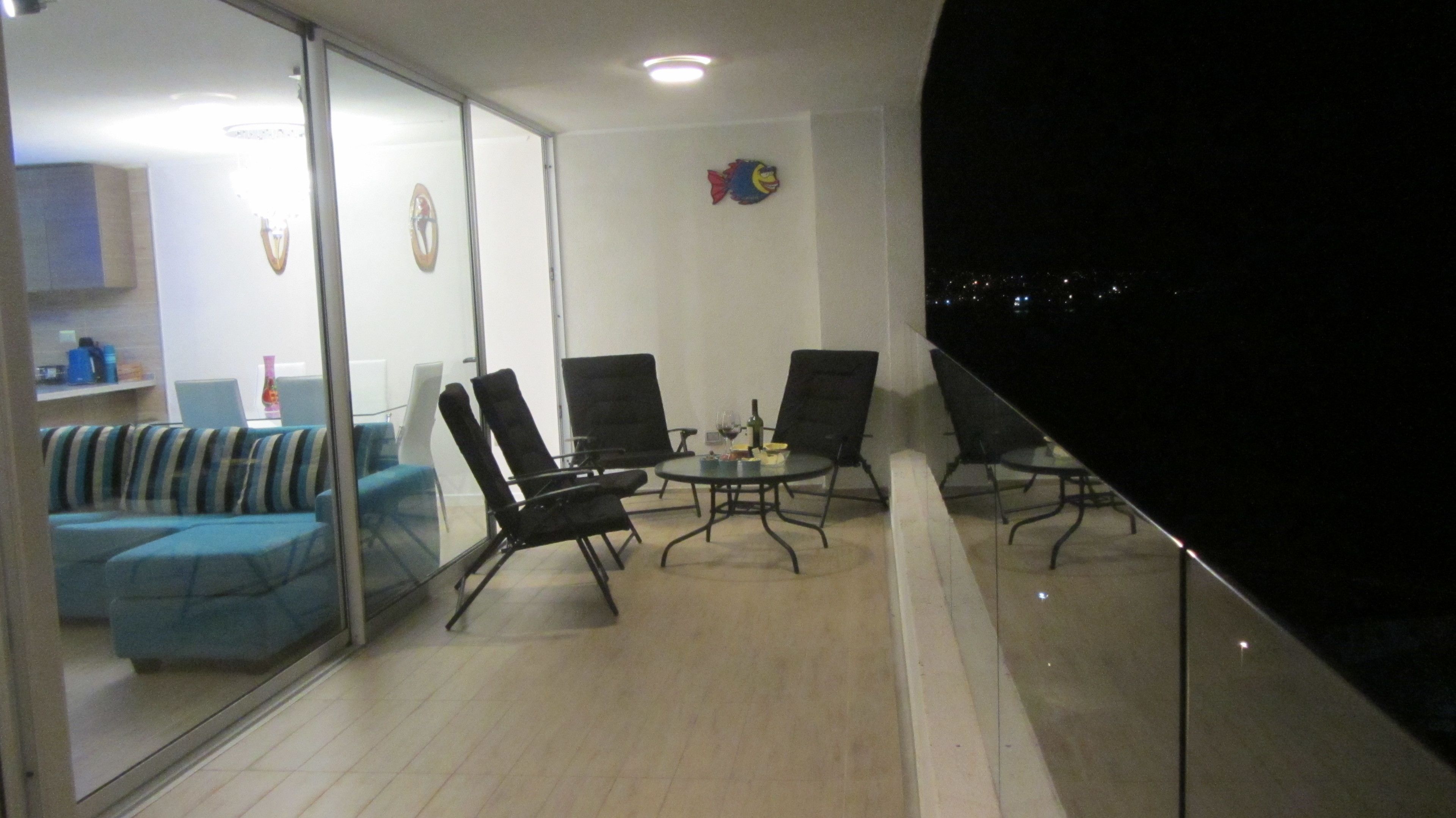 A balcony with a glass partition, two black chairs, a small round table with a bottle and glass, and a sofa with striped cushions.