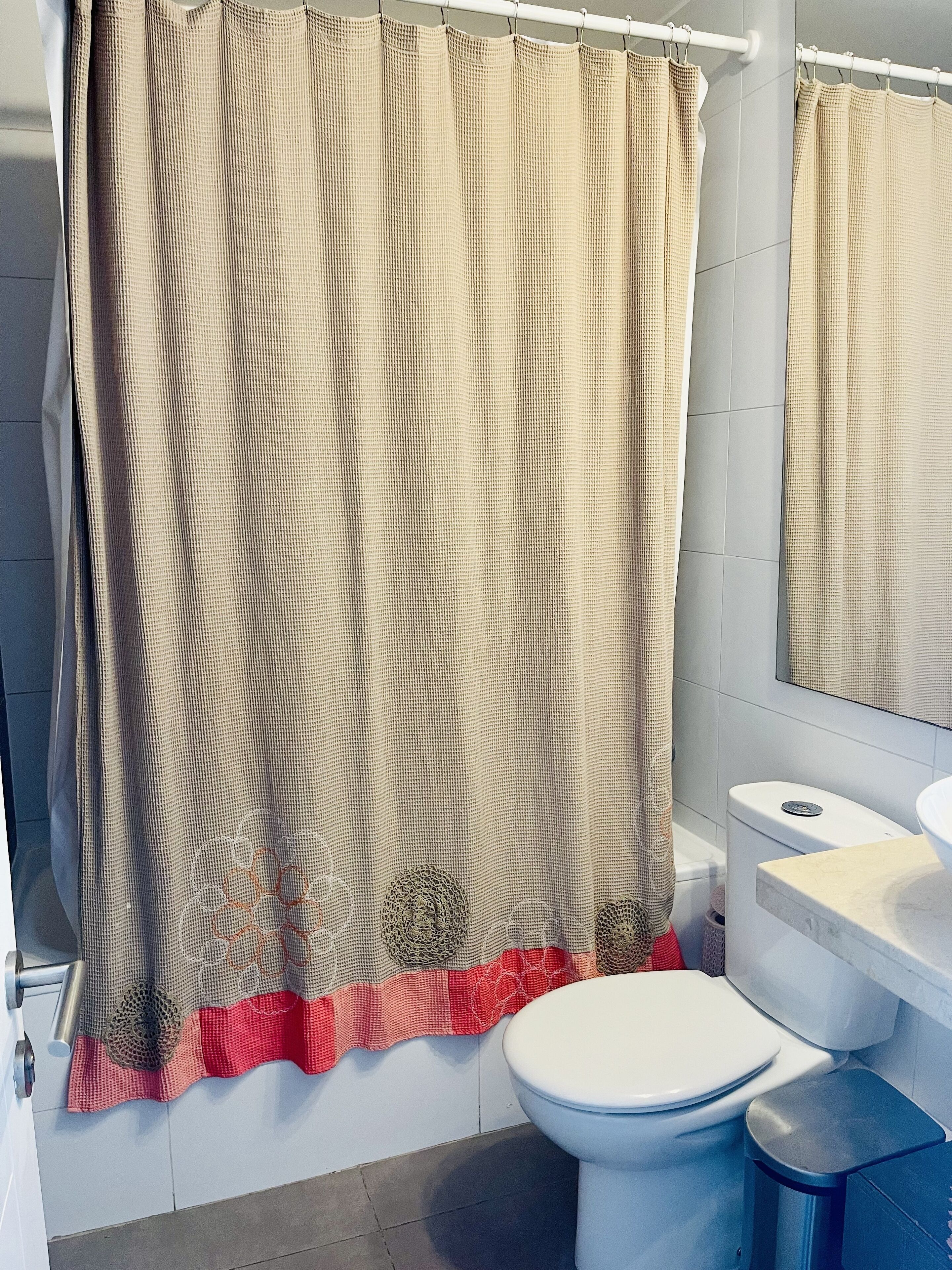 Un baño con cortina de ducha color beige, inodoro blanco y lavamanos con espejo.